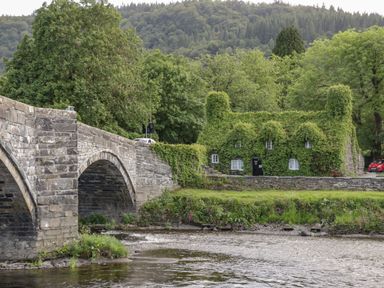 A stone bridge over a river next to a house covered in green ivy with trees and hills in the background at Royal Oak Farm Cottage in Betws-y-Coed