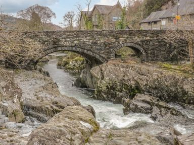 A stone arch bridge over a flowing river with rocks and buildings in the background at Royal Oak Farm Cottage in Betws-y-Coed