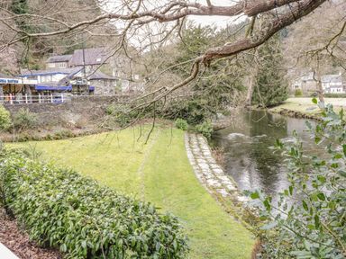 A grassy riverbank with bushes and a stone pathway near a river and buildings in the background at Royal Oak Farm Cottage in Betws-y-Coed
