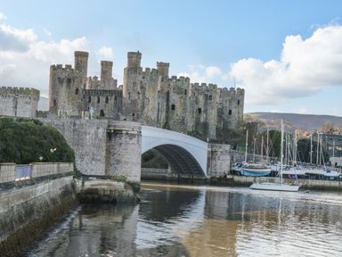 A stone castle with multiple towers next to a bridge over a river with boats docked nearby at Royal Oak Farm Cottage in Betws-y-Coed