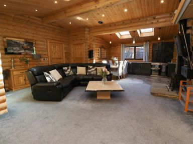 A living area with a black sectional sofa and pillows a square coffee table with flowers a dining table with six chairs a kitchen area with cabinets and stove wooden log walls and ceiling with skylights at Starwood Felton