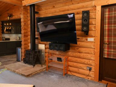 A wall-mounted flat screen TV with speakers and a wood-burning stove next to a kitchen in a log cabin at Starwood in Felton