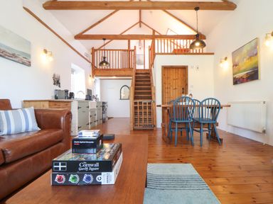 A living area with a brown leather sofa, a wooden coffee table with games on it, a dining table with blue chairs, and a wooden staircase leading to a loft at Joe's Barn in Mullion