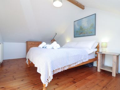 A bedroom with a wooden bed covered by a white patterned blanket and white pillows a wooden side table with a lamp a wooden chest of drawers and a painting on the wall at Joe's Barn in Mullion