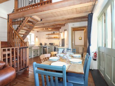 A dining area with a wooden table set for six with plates and glasses next to a kitchen with green cabinets and wooden beams at Berts Barn in Mullion