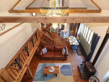A living area with a brown leather sofa a wooden dining table set with plates a wood stove a wooden staircase and a chandelier at Berts Barn in Mullion