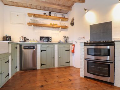 A kitchen with wooden floor green cabinets stainless steel dishwasher double oven microwave and shelves with glassware at Berts Barn in Mullion