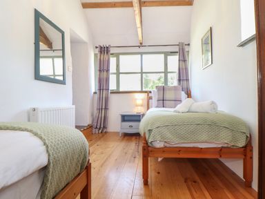 A bedroom with two single wooden beds green bedspreads a bedside table with a lamp and a window with purple curtains at Berts Barn in Mullion