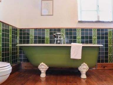 A bathroom with a green clawfoot bathtub a white towel on the tub dark green and patterned tiles on the wall a window and wooden floor at Berts Barn Mullion