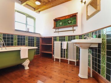 A bathroom with a green clawfoot bathtub a white pedestal sink wooden shelves radiator and window at Berts Barn in Mullion