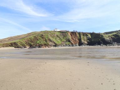 A sandy beach with shallow water in front of a green hill with a house on top at Berts Barn in Mullion