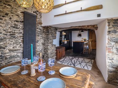 A dining area with a wooden table set with plates and glasses next to a stone wall and an open passage to a kitchen with a wooden island and stools at Pengarreg in Barmouth