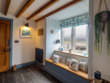 A window seat with striped and bird-themed cushions next to a wooden door and a hanging plant at Pengarreg in Barmouth
