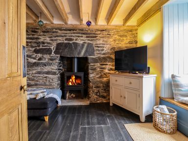 A room with a stone wall fireplace a black wood stove a TV on a white cabinet a blue sofa with blankets and a window seat with cushions at Pengarreg in Barmouth