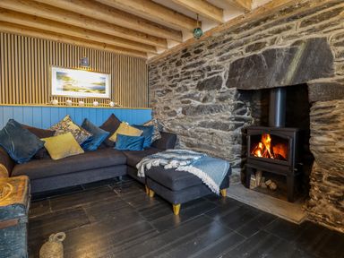 A living room with a dark sofa, a stone wall with a wood burning stove, wooden ceiling beams, and a framed picture on the wall at Pengarreg in Barmouth