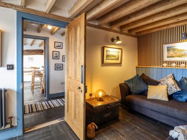 A sitting area with a brown sofa and pillows next to an open wooden door leading to a hallway with wooden chairs at Pengarreg in Barmouth