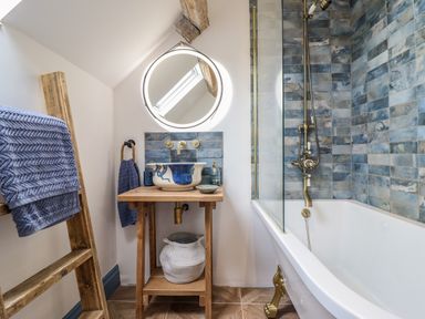 A bathroom with a wooden ladder holding blue towels a wooden sink stand with a blue and white basin a round mirror above the sink and a bathtub with a shower and blue tiled wall at Pengarreg in Barmouth