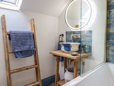 A bathroom with a wooden ladder holding blue towels a wooden sink stand with a ceramic basin a round mirror and a bathtub with a glass screen at Pengarreg in Barmouth