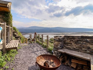 An outdoor seating area with wooden benches a fire pit stone walls and a view of hills and water at Pengarreg in Barmouth