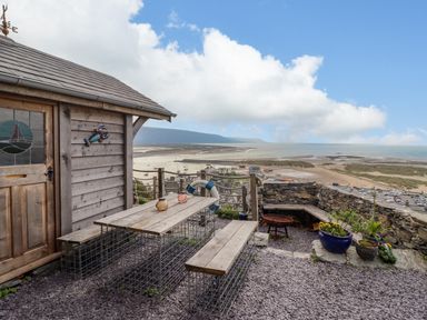 An outdoor seating area with a wooden table and benches next to a small wooden shed overlooking a beach and sea at Pengarreg in Barmouth
