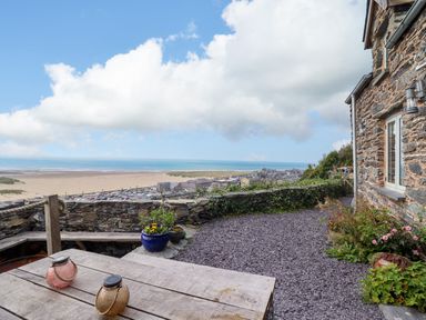 An outdoor patio area with a wooden table, stone wall, potted plants, and a beach view at Pengarreg in Barmouth