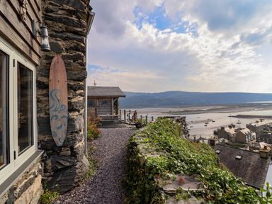 A stone house patio with a wooden surfboard decoration a gravel path and a view of the sea and rooftops at Pengarreg in Barmouth