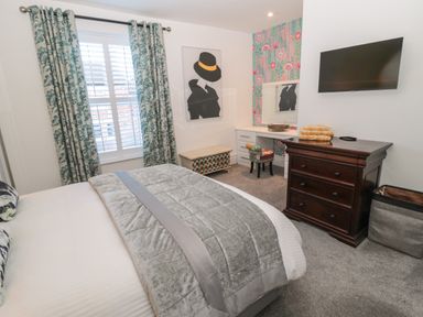 A bedroom with a bed, window with curtains, chest of drawers, dressing table with a chair, and wall art at Sunflowers in Earsdon near Whitley Bay