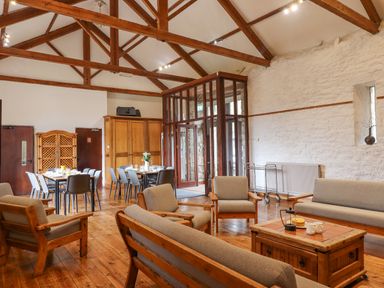 A sitting area with wooden sofas and a coffee table next to dining tables and chairs in a room with exposed wooden beams and white stone walls at Brimpts Barn Dartmeet near Princetown