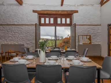 A dining room with a set table facing open wooden doors leading to a balcony with a view of trees at Brimpts Barn in Dartmeet near Princetown