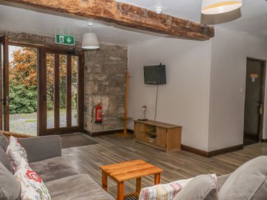 A sitting area with a gray sofa pillows a small wooden table a TV on a wooden stand stone wall and wooden floor at Brimpts Barn Dartmeet near Princetown