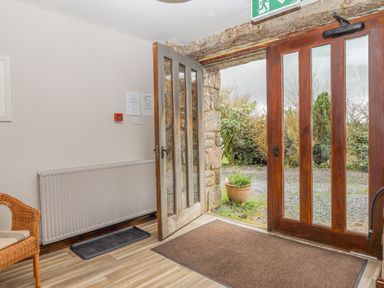 An entrance area with an open wooden door, a large doormat, wicker chair, radiator, and a view of a garden at Brimpts Barn in Dartmeet near Princetown