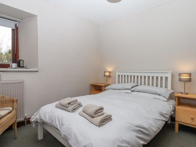 A bedroom with a double bed with folded towels bedside tables with lamps a wicker chair and a window at Brimpts Barn in Dartmeet near Princetown