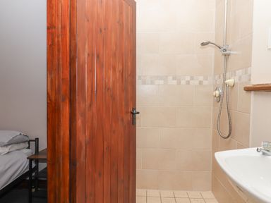A shower area with beige tiles and a wall-mounted showerhead next to a white sink with a faucet and a partially visible wooden door with bedroom furniture behind it at Brimpts Barn Dartmeet near Princetown