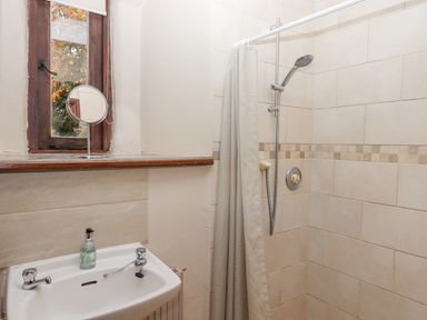 A bathroom with a shower area, a shower curtain, a sink with soap dispenser, and a window with a round mirror on the sill at Brimpts Barn Dartmeet near Princetown