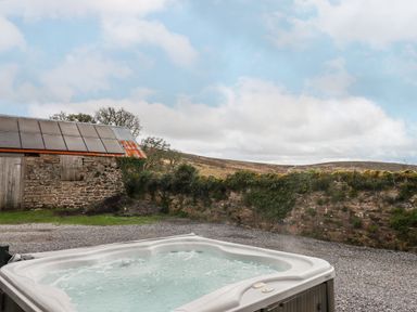 An outdoor hot tub on a gravel area near a stone wall and a building with a corrugated metal roof at Brimpts Barn Dartmeet near Princetown