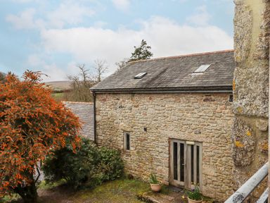 A stone building with a slate roof and double doors surrounded by bushes and trees at Brimpts Barn in Dartmeet near Princetown