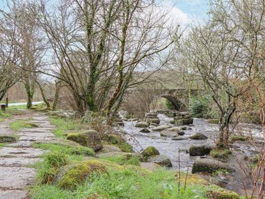 A stone path beside a river with rocks and trees and a stone bridge in the background at Brimpts Barn Dartmeet near Princetown