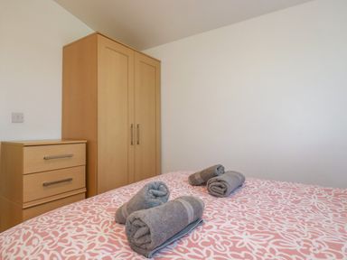 A bedroom with a bed covered in a patterned blanket and four rolled towels on top next to a wooden wardrobe and chest of drawers at Cornish Holiday at Atlantic Bays in St Merryn