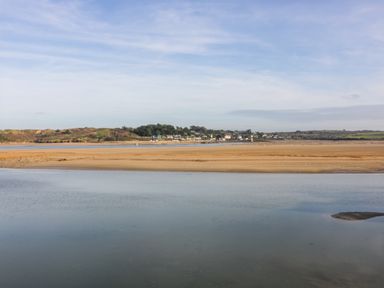 A coastal landscape with water in the foreground and a sandy shore with houses and hills in the background at Cornish Holiday at Atlantic Bays in St Merryn