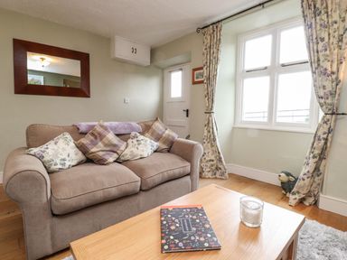 A living room with a beige sofa with patterned cushions a wooden coffee table with a candle and a visitors book floral curtains and a window at Lavender Cottage in Coniston