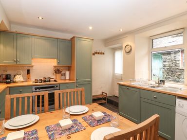 A kitchen with green cabinets a wooden dining table set with plates and glasses and a window showing a stone wall at Lavender Cottage in Coniston