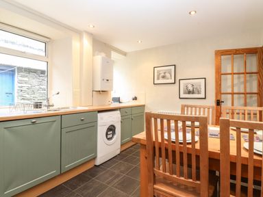 A kitchen with green cabinets a washing machine a wooden dining table with chairs and framed pictures on the wall at Lavender Cottage in Coniston