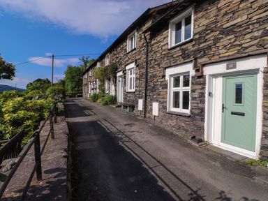 A row of stone cottages with white framed windows and a green door along a paved path with plants and a metal railing at Lavender Cottage in Coniston