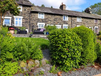 A stone house with white framed windows and a garden with bushes and plants at Lavender Cottage in Coniston