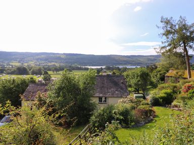 A house with a slate roof surrounded by trees and bushes with fields and hills in the background at Lavender Cottage in Coniston