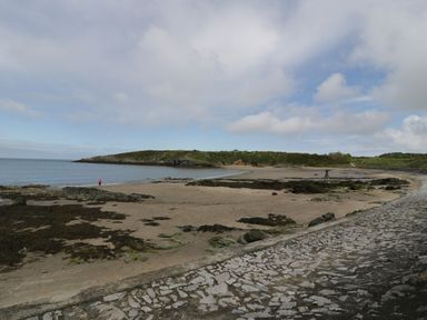 A sandy beach with rocks and seaweed near a stone paved walkway and a person standing by the shoreline with a green hill in the background at Ty Bara in Cemaes Bay