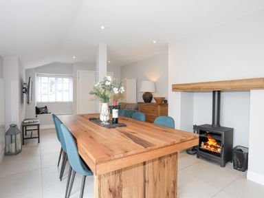 A dining room with a wooden table blue chairs a vase with flowers a bottle of wine a wood burning stove and a chest of drawers at Ty Bara in Cemaes Bay