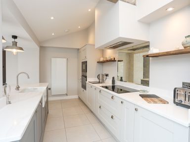 A kitchen with white countertops gray cabinets a sink a stove a toaster and wooden shelves at Ty Bara in Cemaes Bay