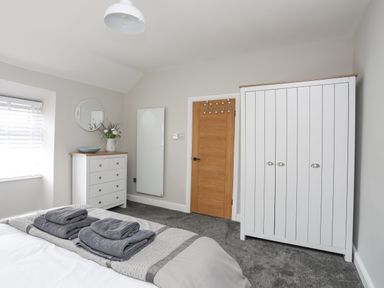 A bedroom with a bed, white dresser with flowers, a mirror, a wooden door, and a white wardrobe at Ty Bara in Cemaes Bay