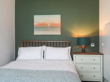 A bedroom with a bed white bedding a wooden headboard a nightstand with a lamp and a painting on a green wall at Ty Bara in Cemaes Bay